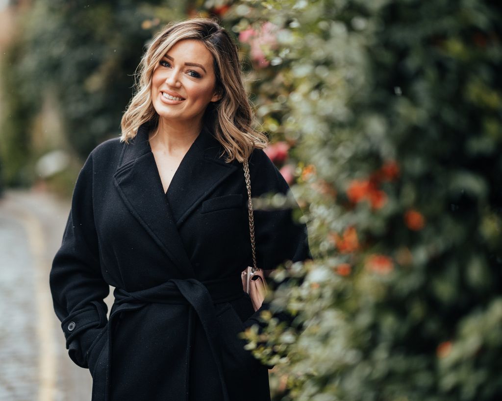 young woman smiling in a black coat standing next to a hedge 